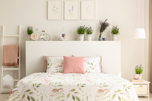 Pink pillow on bed with headboard in white bedroom interior with plants and posters. Real photo