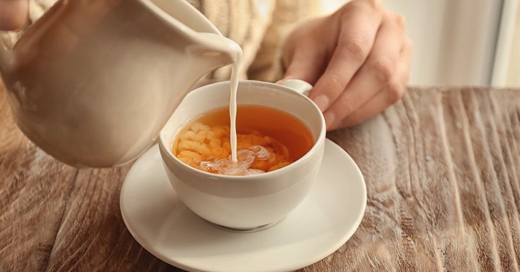 Xícara com chá branca e pires branco em mesa. Mãos colocando leite de uma leiteira no chá.
