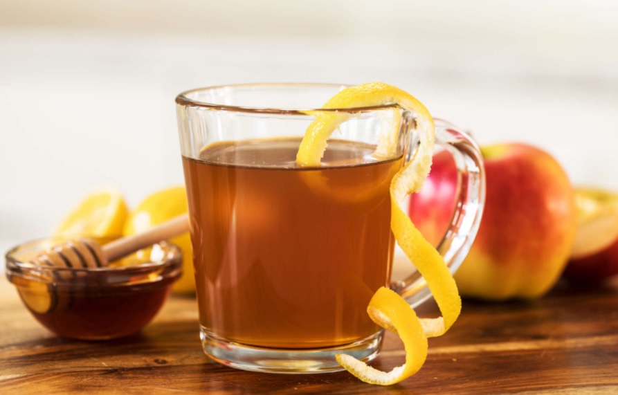Xícara de vidro de chá de maçã. Casca de limão siciliano enrolada pendendo da xícara. Maçãs e pote com mel ao fundo