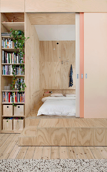 Gif de quarto com piso e parede em madeira. Porta de correr em tons de rosa abrindo e fechando revelando a cama. Estante com livros e planta