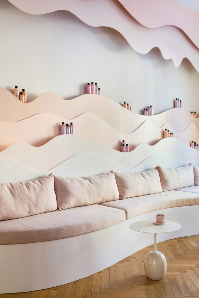 Sofá com almofadas rosa claro. Enfeites na parede e prateleiras em forma ondulada com produtos