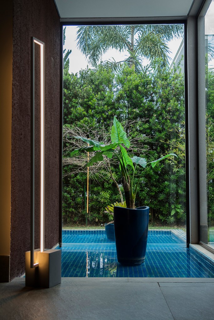 Luminária minimalista ao lado de uma planta grande e uma parede de vidro com vista para o quintal