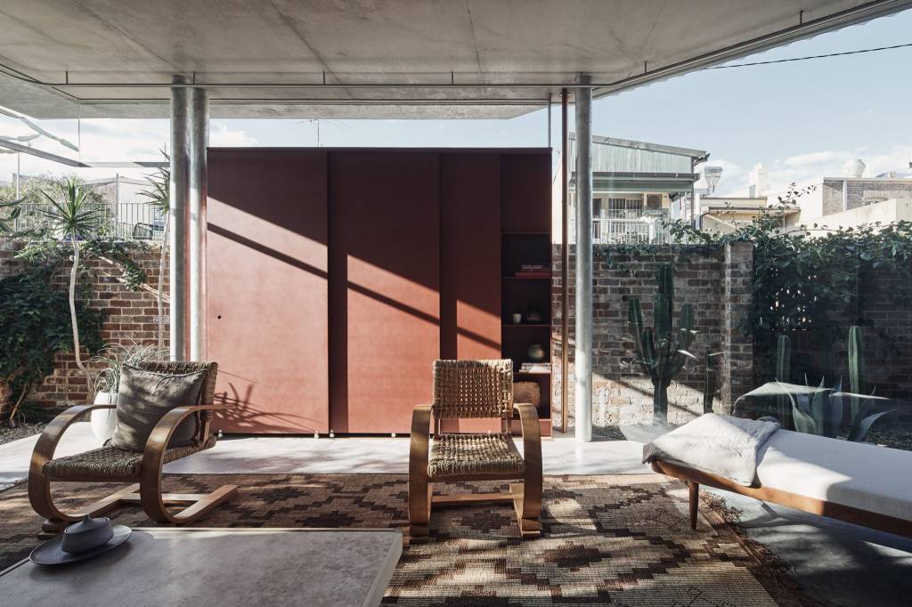 Sala com paredes de vidro, com colunas de metal. A mobília tem o mesmo estilo descontraído, com cadeiras de madeira e um sofá-cama, criando locais de descanso confortáveis.