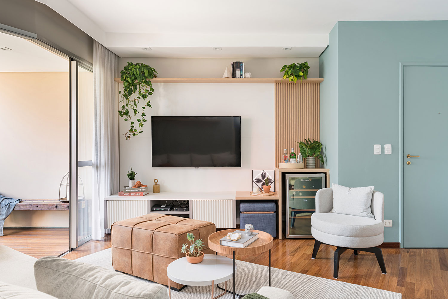 Sala de estar com piso de madeira, marcenaria azul, banco branco, televisão fixa na parede e plantas trepadeiras