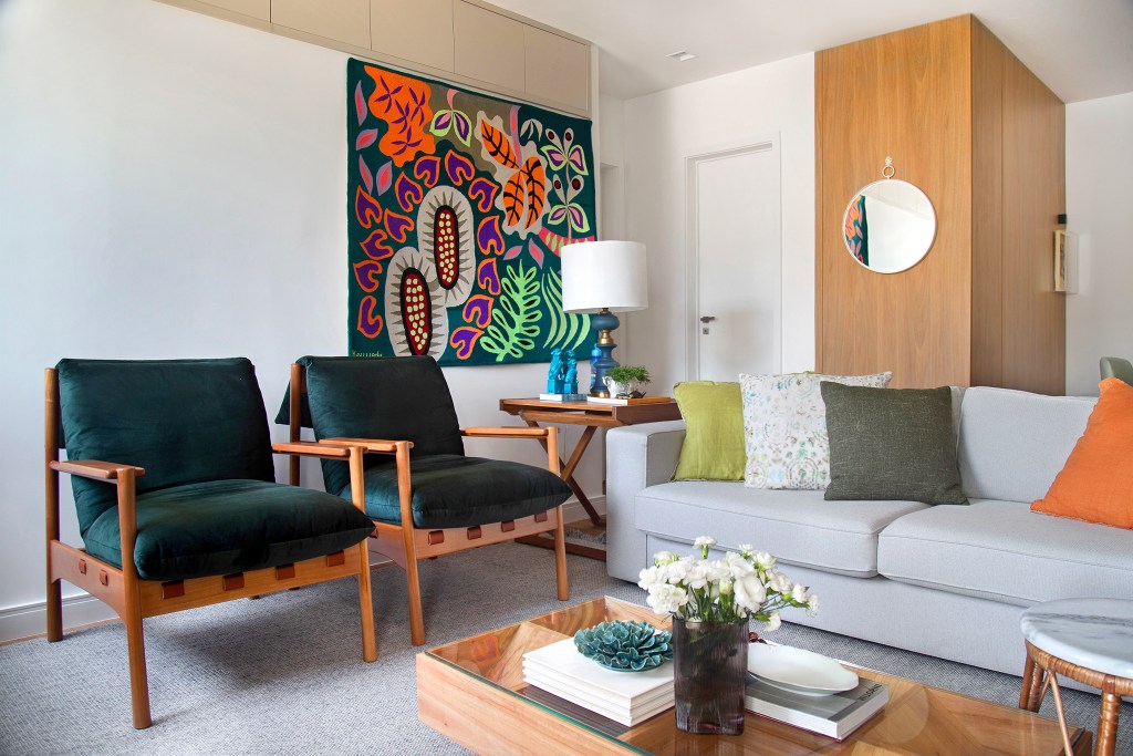 Sala de estar com tapeçaria colorida na parede, duas poltronas verdes, um sofá cinza claro e uma mesa de centro de madeira