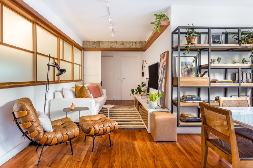 Sala de estar com piso de madeira e décor neutro, estante no estilo industrial, sofá branco e poltrona marrom