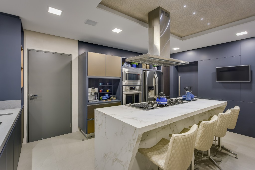 Cozinha com armários azul escuro pastel, e eletrodomésticos em inox. No centro, uma ilha com cooktop em pedra branca, com cadeiras na mesma cor