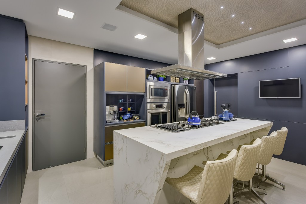 5-dicas-para-a-cozinha-perfeita-casa.com-1 2017©Nenad-AlessandraGandolfi-2101-2-EditarAlta Cozinha com armários azul escuro pastel, e eletrodomésticos em inox. No centro, uma ilha com cooktop em pedra branca, com cadeiras na mesma cor
