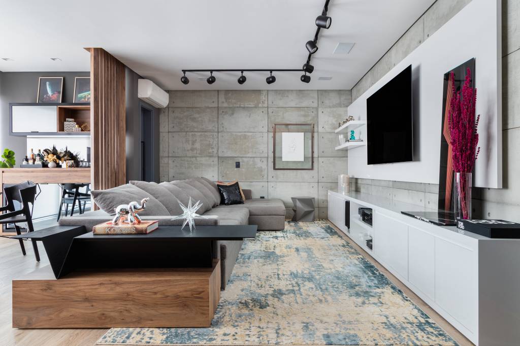 Sala de estar da Casa Vila Mariana, com moveis brancos, cinza e madeira. No chão, porcelanato e na parede, revestimento de cimento queimado