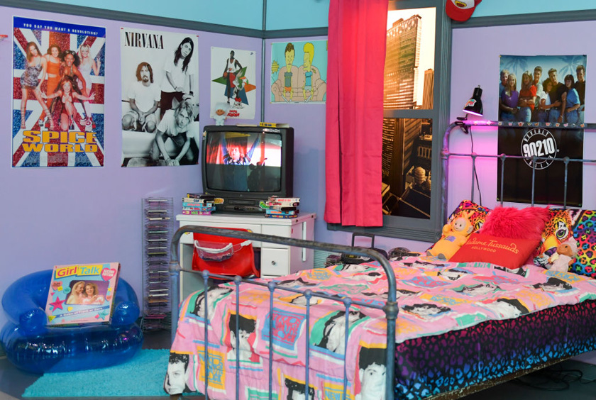 Madame Tussauds Hollywood Welcomes Tom Green To Unveil New ’90s Room Launch Quarto com paredes lilás. Cama com colcha estampada. Posteres na parede. Puf azul inflável. Televisão de tubo