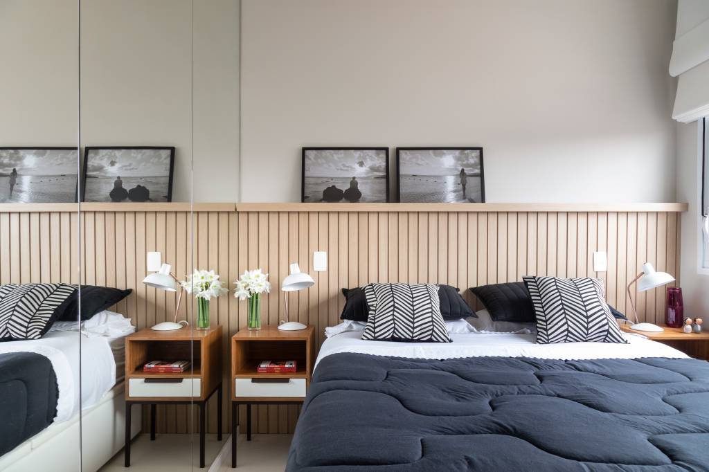 Quarto do casal, com cabeceira de madeira ripada, doi quadros em preto e branco acima dela. Na lateral da cama, uma mesa de madeira com uma gaveta branca, livros no nicho pequeno e flores sobre ela. Na cama, o edredom é azul escuro. Na lateral, um espelho de fora a fora esconde o guarda roupas