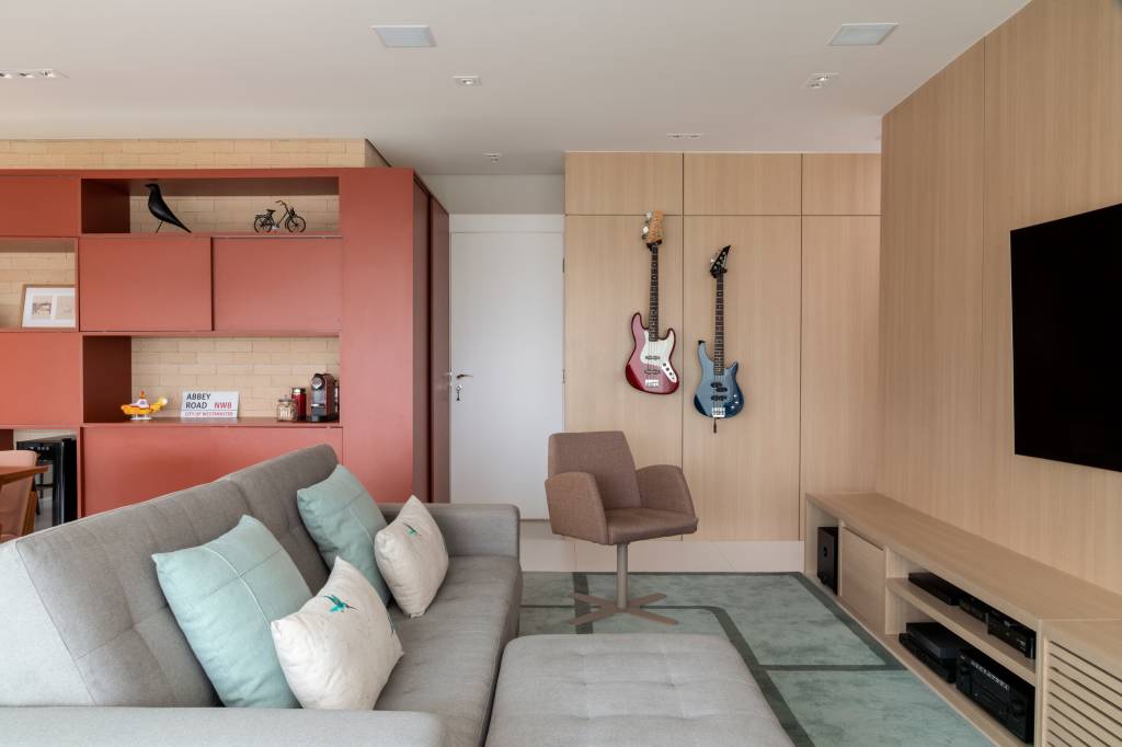 Sala de estar com marcenaria na parede, com duas guitarras presas a ela em suportes. O sofá cinza é adornado com duas almofadas verde claro e duas brancas. No chão, um tapete verde. Ao fundo, do lado esquerdo, armário na cor goiaba