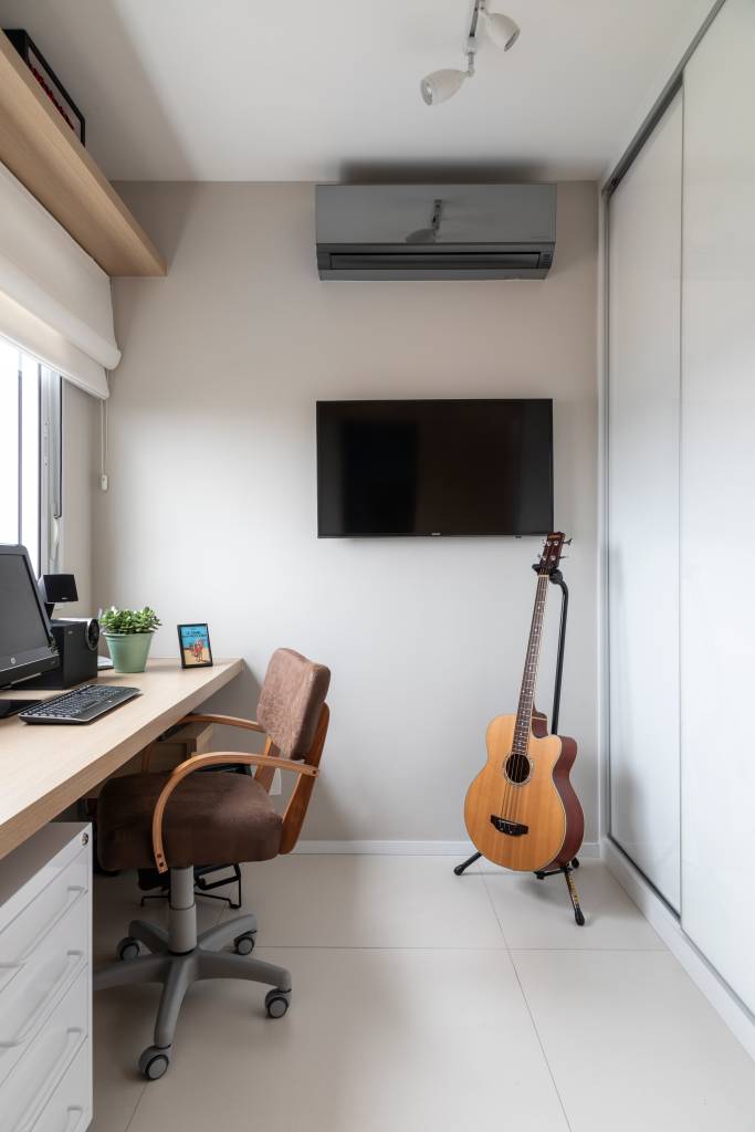 escritório constituído por bancada, cadeira ergonômica, móveis soltos e iluminação natural. No canto, uma Tv e um violão em pé no suporte