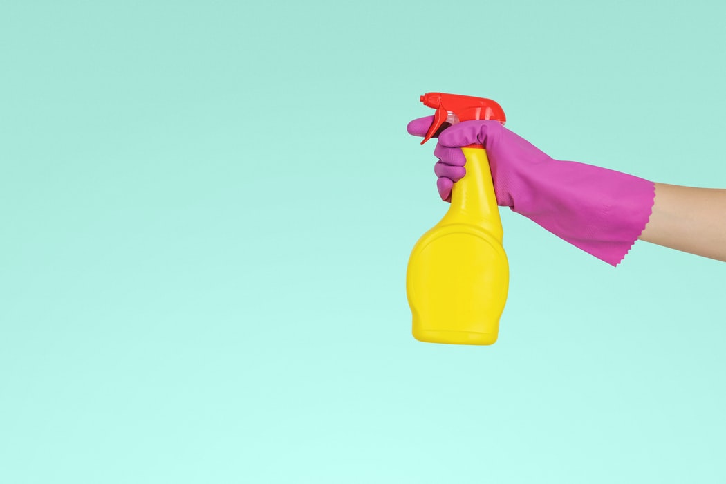 Fundo azul com um mão com luva rosa segurando uma embalagem de spray amarela