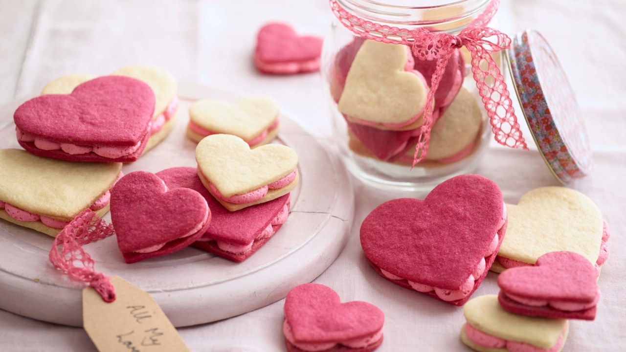 Biscoitos em formato de coração amarelos e rosas. Pote de vidro com fita rosa e mais biscoitos dentro ao fundo