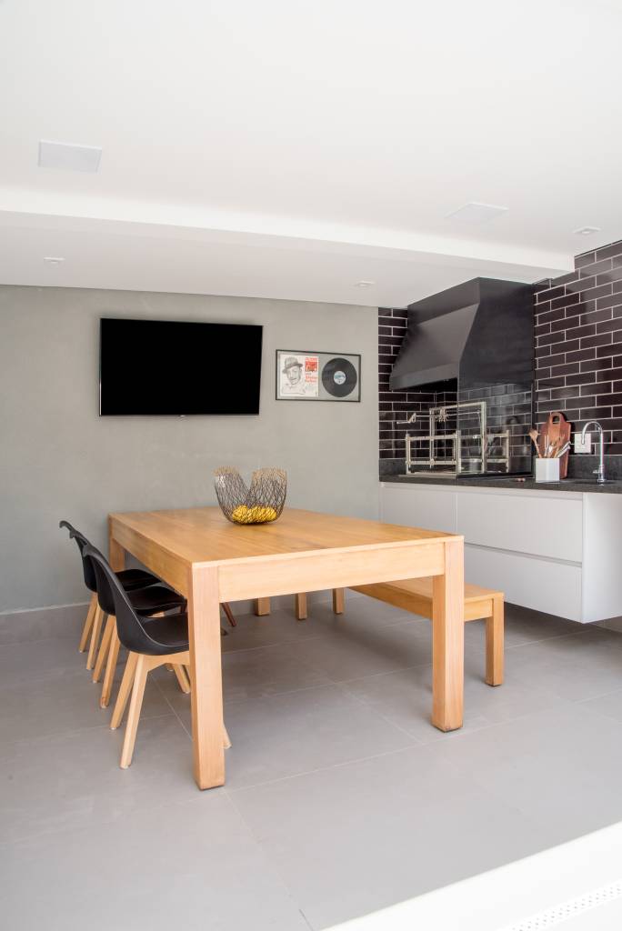 revestimentos-para-varanda-casa.com-Julia-Herman-5 Área gourmet, com paredes cinzas e TV pendurada em uma delas, área da churrasqueria com revestimento de tijolinhos pretos, com bancada branca, mesa de madeira com um banco no mesmo material e três cadeiras com assentos pretos