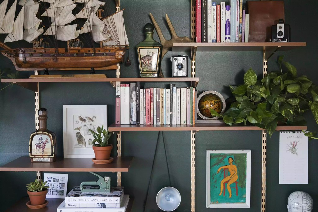 trends-anos-90-the-spruce-casa.com-5-jessica-brigham Estante de madeira, com prateleiras presas por cordas em alturas irregulares, com fundo vazado para a parede verde esmeralda. Livros, quadros e itens decorativos estão na estante