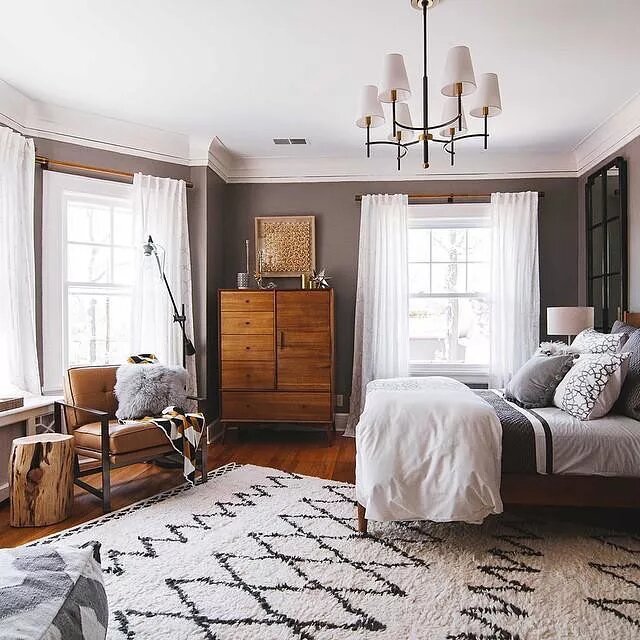 trends-anos-90-the-spruce-casa.com-8-west-elm Quarto com paredes cinzas e móveis de madeira escura. No chão um tapete branco com padrão geométrico. A cama está do lado direto, com muitos travesseiros, sobre um lustre