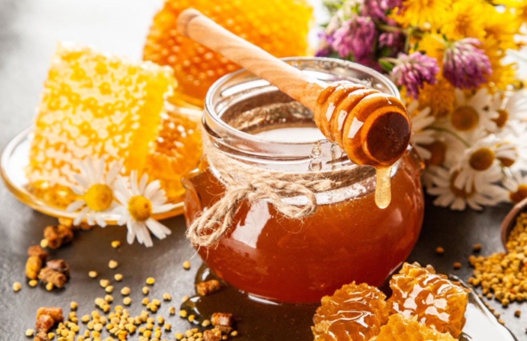 pote de mel de vidro arredondado com colher de pau coberta de mel. favas e flores espalhadas ao fundo