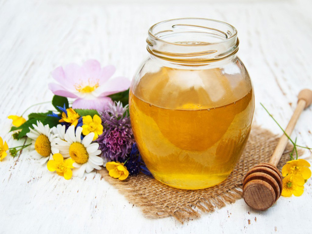 pote de mel arredondado de vidro em mesa de madeira branca com pequenas flores ao lado