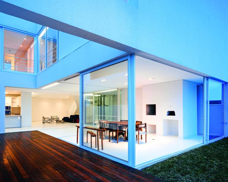 Quando a tarde cai, o espaço a céu aberto do deck traz a luz natural até os quartos do primeiro andar (no alto, à esq.) e surge tingido de azul. No térreo, a ausência de colunas eo cumaru do solário nivelado ao piso de resina epóxi garantem o entrelaçamento dos espaços.
