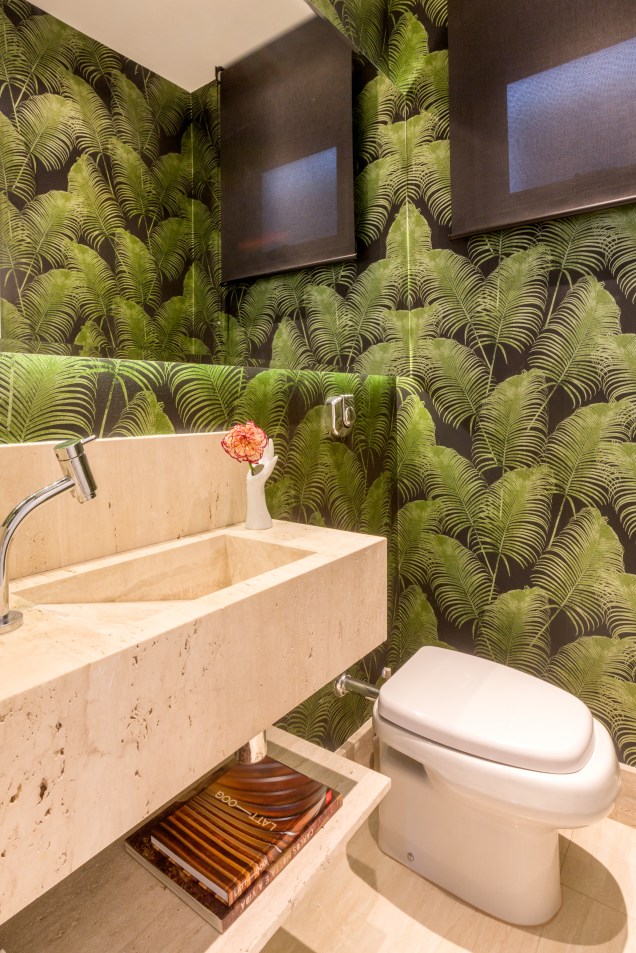 O toque praiano do lavabo fica por conta do papel de parede (Wallcovering)  com folhas verdes e fundo escuro colocado em todas as paredes. A bancada com pia esculpida e prateleira baixa é feita de Mármore Travertino.