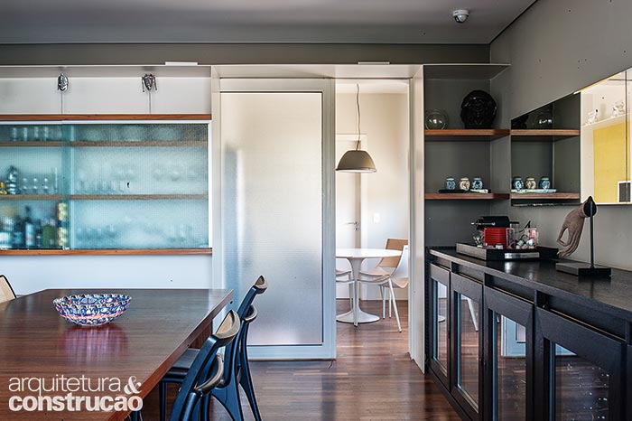 De chapa de ferro pintada de branco, a estante percorre o apartamento e se ajusta às necessidades de cada ambiente. Na sala de jantar, desenha a cristaleira e um pórtico, atrás do qual se esconde a porta, também de ferro. O vidro aramado garante luz e privacidade. Projeto de Pessoa Arquitetos e Base Urbana.