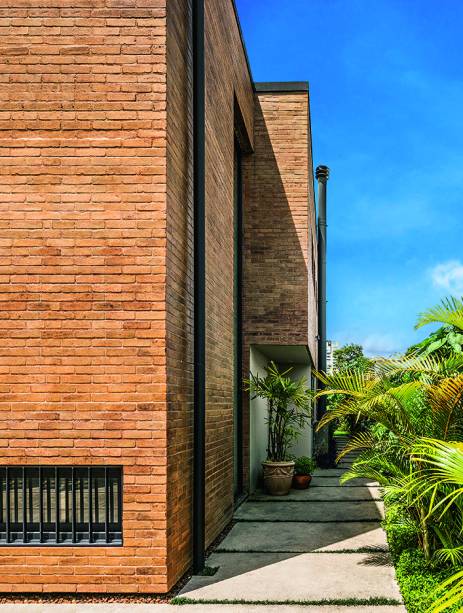 Por todo lado (esta é a vista que se tem ao ultrapassar o portão da garagem), a cor cinza-chumbo confere unidade a elementos distintos: os tubos de água pluvial (aparentes, para facilitar a manutenção), a chaminé da lareira, o acabamento da platibanda, guarda--corpos e gradis. O mesmo tom aparecenas esquadrias de PVC (Eurosystem). CAPRICHO ARTESANAL:  O tijolinho da fachada é o tradicional, de barro, partido ao meio no sentido do comprimento. A escolha levou em conta o melhor acabamento nas quinas em relação às opções vendidas em forma de plaqueta