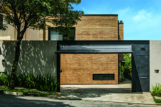Desde a entrada, partes metálicas tingidas em tom escuro reforçam a geometria da construção e contribuem para um resultado discreto. É o caso do portão da garagem, uma ampla superfície cinzenta. Mais adiante, lá dentro, a janela baixa e longilínea (dotada de grades de ferro) fica diante da escada para o subsolo, recortando a face no geral bastante fechada para a rua.
