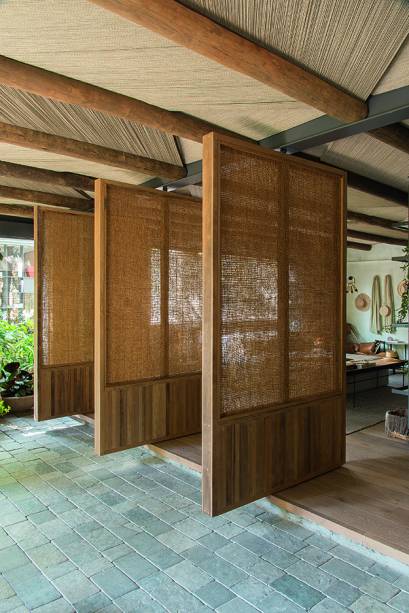 Em comemoração aos dez anos de seu escritório, o Triplex Arquitetura, o trio Adriana A. Helú Hawilla, Carolina Oliveira e Marina Torre Lobo concebeu a Casa Raízes. No espaço de 115 m², pedra, madeira e palha são os elementos eleitos. O projeto também valorizou iluminação e ventilação naturais, reflexo do pensamento sustentável das sócias. “Queríamos integração entre fora e dentro, por isso derrubamos uma parede existente entre o estar e a varanda e instalamos as portas pivotantes”, diz Adriana. Executadas pela Hydrotech, elas abraçam pilares de sustentação, camuflando parte da estrutura metálica de reforço. Confecção manual: No forro, vigas de eucalipto (Hydrotech) sustentam as placas com tramas (Nani Chinellatto) feitas fio a fio, formando o desenho geométrico.