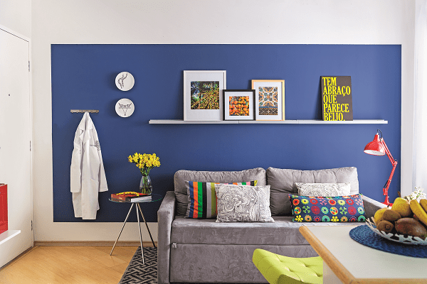 <span>A estrela da sala é a parede azul – note que ela não é inteiramente tingida, funcionando como um painel pintado. Neto dá as dicas para copiar: “A referência para a altura deve ser a porta ou uma viga, o que estiver mais visível para equilibrar o alinhamento. Já para as laterais e a parte de baixo, sugiro afastamento de 15 a 20 cm”. A proposta valoriza a superfície sem deixar o ambiente pesado. “Uma saída para quem tem medo de abusar do tom escuro”, aponta o arquiteto. Tintas da sala: painel azul cor Tratamento Real e demais paredes cor Cinza de Grife, da Coral. Acrílico fosco. </span>Projeto do arquiteto Neto Porpino