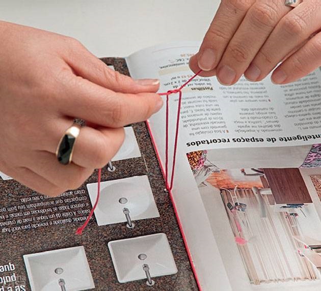 16. Ali, você encontrará a extremidade da linha (deixada no passo 12), que deverá ser atada a essa da agulha. Antes, passe uma delas por baixo do trecho do fio que une os furos de cima e de baixo para que o nó o abrace. Dê dois nós e corte. Repita os passos (a partir do 11º) com as outras revistas.