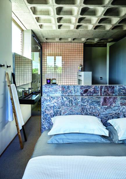 Da entrada do quarto do casal, é possível avistar as instalações do banheiro, onde a decoração focou em acessórios elegantes. A única delimitação entre os ambientes é a cabeceira de mármore português, em placas de 30 x 30 cm.