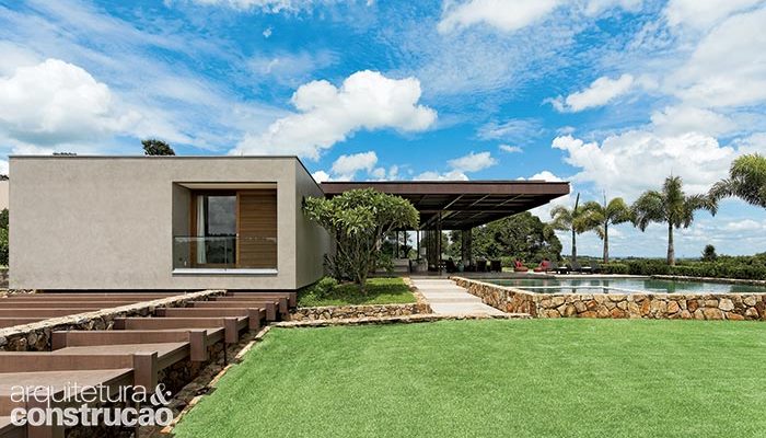 A casa está posicionada no platô encontrado no terreno. À esq. nesta foto, vê-se o fechamento superior da garagem, a qual conta com um longo corredor de entrada escavado num nível abaixo do volume térreo. Na fachada, textura de base acrílica da Terracor (ref. Cimento Queimado).