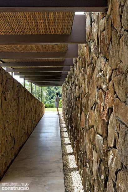 Entrada da casa: escavada terreno adentro, é vizinha da garagem e conta com pérgula de aço e forro de palha. Nas paredes laterais, pedra moledo.
