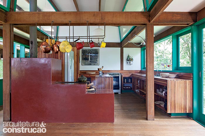 Marcando os limites da cozinha, está o fogão a lenha revestido do tradicional vermelhão. As panelas penduram- se na armação de cobre presa às vigas. Bancadas e armários de eucalipto laminado.