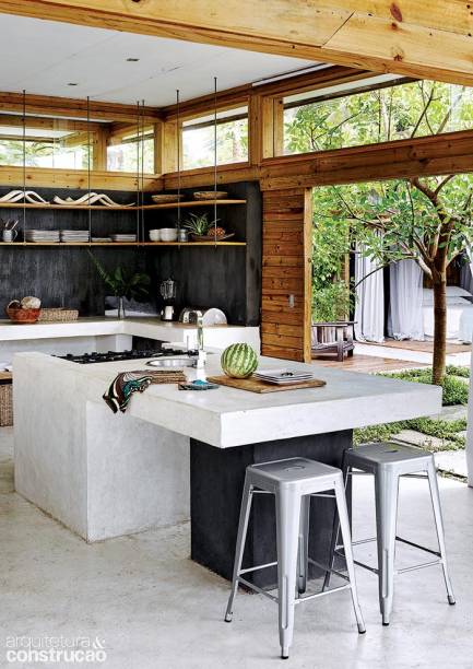 Atrás da bancada de concreto da cozinha, a parede escura leva cimento cinza com pigmento preto e gesso para dar liga. A textura vem da porção de areia adicionada à mistura na própria obra. Prateleiras suspensas de madeira acomodam as louças.