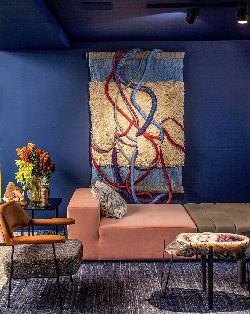 Parede azul com tapeçaria pendurada, em frente a uma namoradeira, uma cadeira e uma mesa lateral.