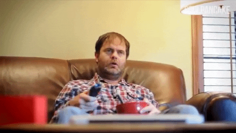 Rainn Wilson sentado num sofá, ligando a tv com cara emburrada