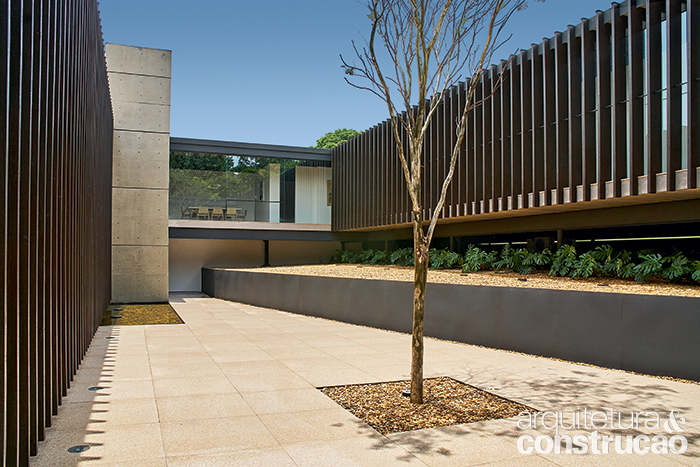 Fonte de luminosidade para o térreo, o pátio resume os materiais usados na casa: piso cimentício com microgrânulos, laje pré-fabricada de concreto, vigas e pilares de metal e fechamentos de vidro e de madeira, esta vista no brise  A cobertura (Omnitrade Brasil) é de manta termoplástica aplicada sobre painéis de OS B com isolante termoacústico.
