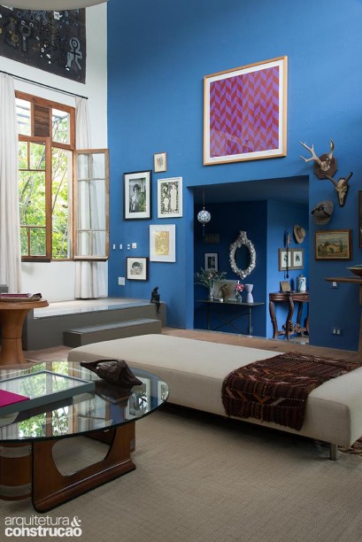 O azul da fachada invade a sala, colorindo a lateral com pé-direito duplo. Nela, encontra-se a passagem para a antiga cocheira.