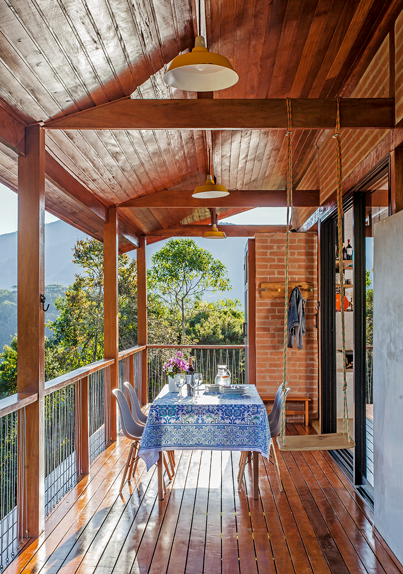 Casa de campo suspensa é prática e teve custo barateado. Na foto, varanda com deck de madeira, mesa com cadeira, árvores ao fundo.