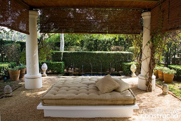 Numa base de alvenaria e sob o pergolado com forro de palha, o futon é o principal ponto de convívio do jardim.