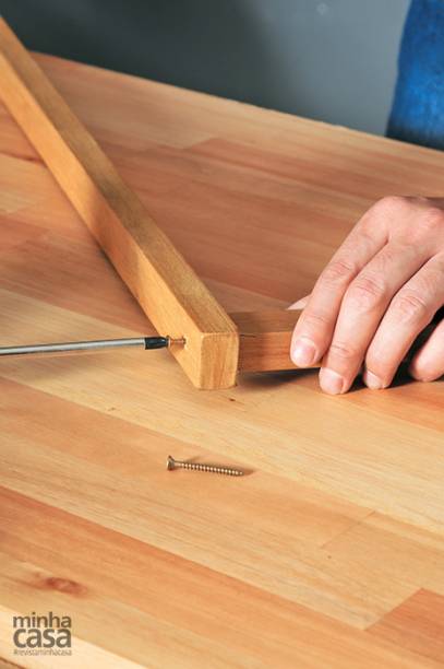 10. Antes que seque, usa a chave de fenda para parafusar esse pedaço, com o lado de 3cm voltado para cima, a um dos furos do sarrafo grande. Segure firme na posição até fixar. Faça o mesmo com o outro furo do sarrafo e repita este passo com as outras duas peças maiores.