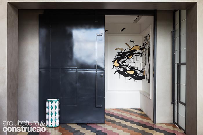 A porta corrediça de aço-carbono (Serralheria Arco Íris) com pontos de solda aparentes e a parede grafitada definem o hall, até onde chegam os ladrilhos hexagonais.