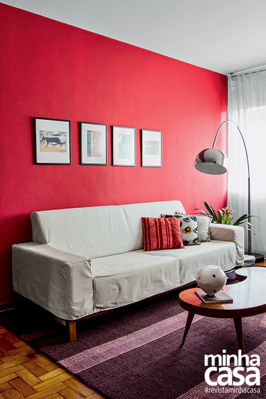 Sala de estar com sofá de capa cinza, parede vermelha e quadros acima do sofá.