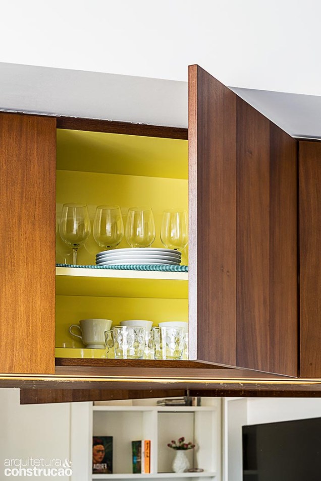 Externamente, o armário superior da cozinha é coberto de lâmina de itaúba escurecida. As portas dispensam puxador, proporcionando maior limpeza visual. Abertas, trazem à tona uma bela surpresa: o toque vibrante do revestimento melamínico amarelo, aplicado na parte de dentro.