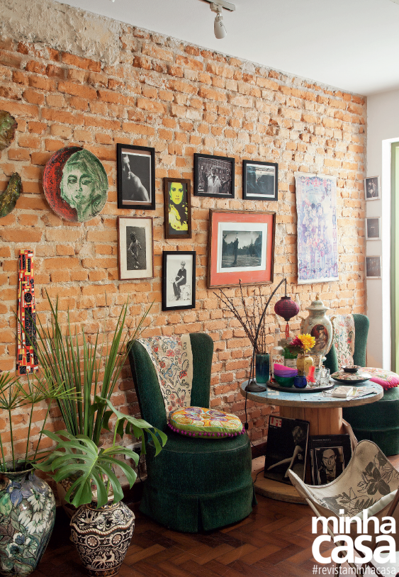 Hall aconchegante, com cadeiras, uma mesinha redonda, plantas no chão, e quadros pendurados em uma parede de tijolos.
