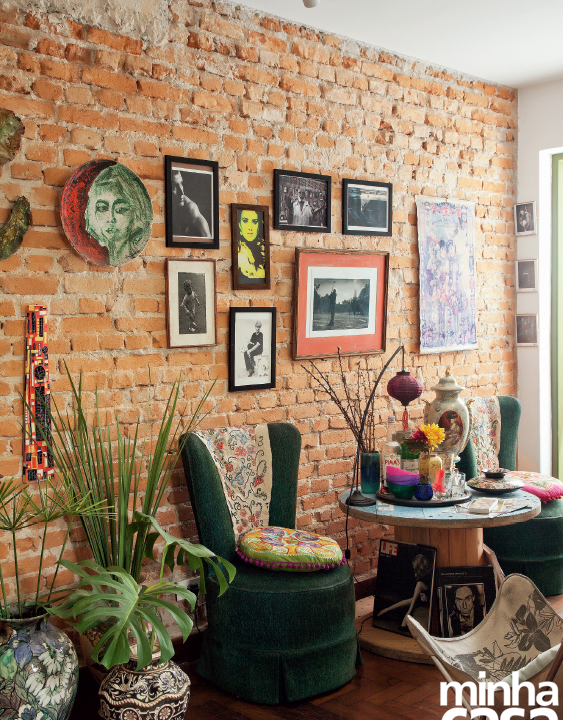 Hall aconchegante, com cadeiras, uma mesinha redonda, plantas no chão, e quadros pendurados em uma parede de tijolos.