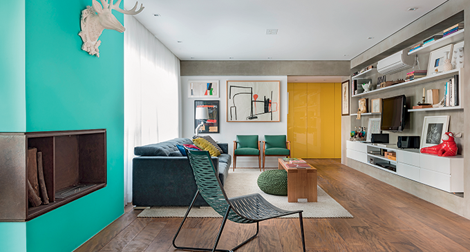 Sala de estar com grande porta amarela, poltronas verdes, quadro colorido com figuras geométricas, sofá azul e uma cadeira de descanso ao lado.